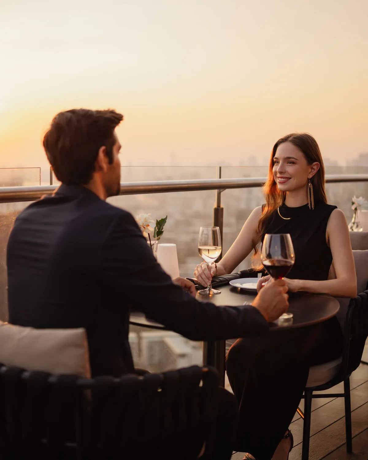 Paar beim Dinner auf einer Dachterrasse bei Sonnenuntergang, die Frau lächelt ihr Gegenüber an