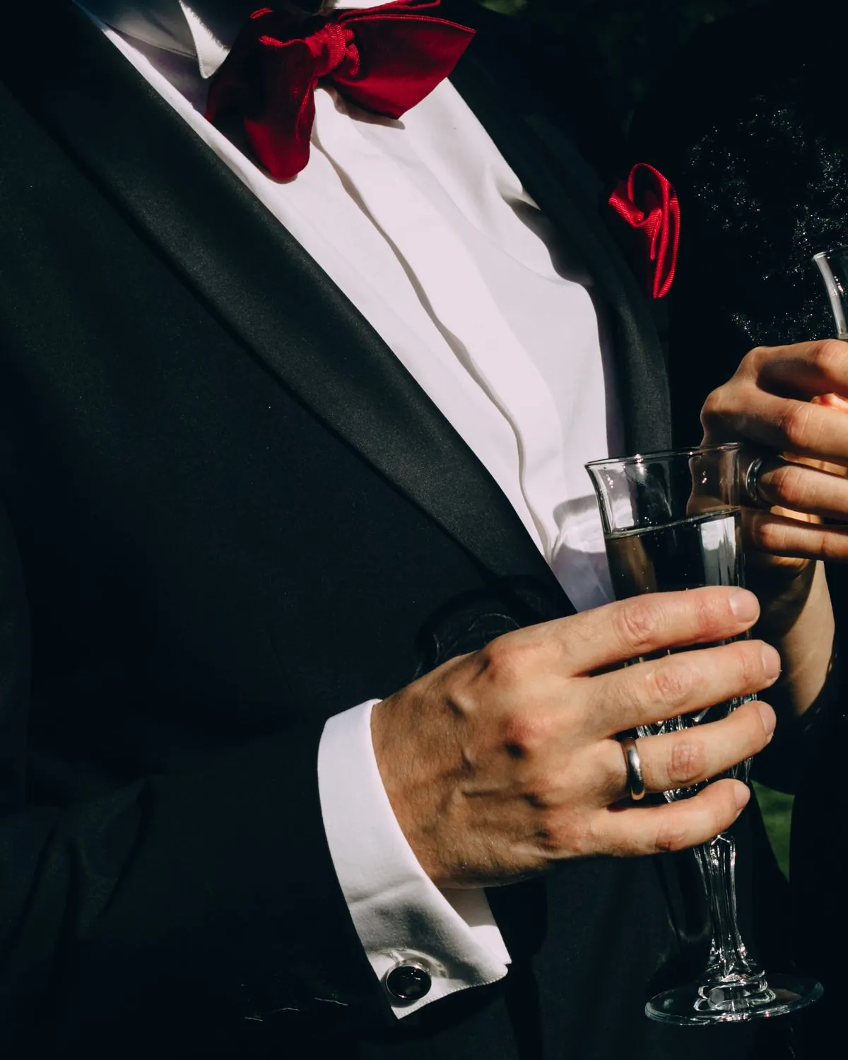 Herr im Smoking mit Sektglas auf einem Abendempfang, daneben die Hand seiner Begleitung mit Glas