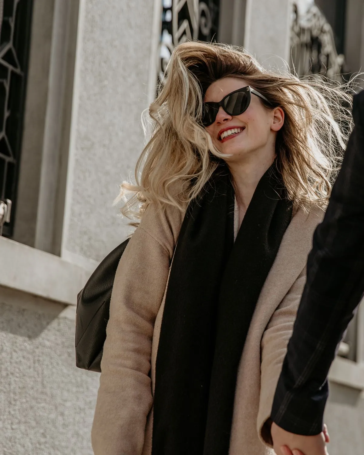 Lachende Frau mit Sonnenbrille und wehendem Haar geht in der Stadt Hand in Hand mit ihrem Begleiter