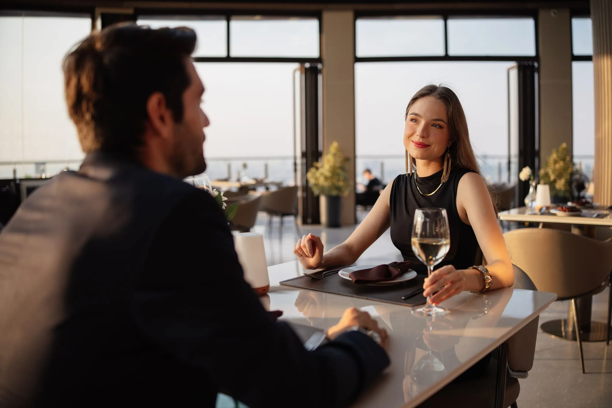 Eine Frau im schwarzen Kleid sitzt beim Dinner mit einem Weinglas am Tisch und lächelt ihrem Gast zu