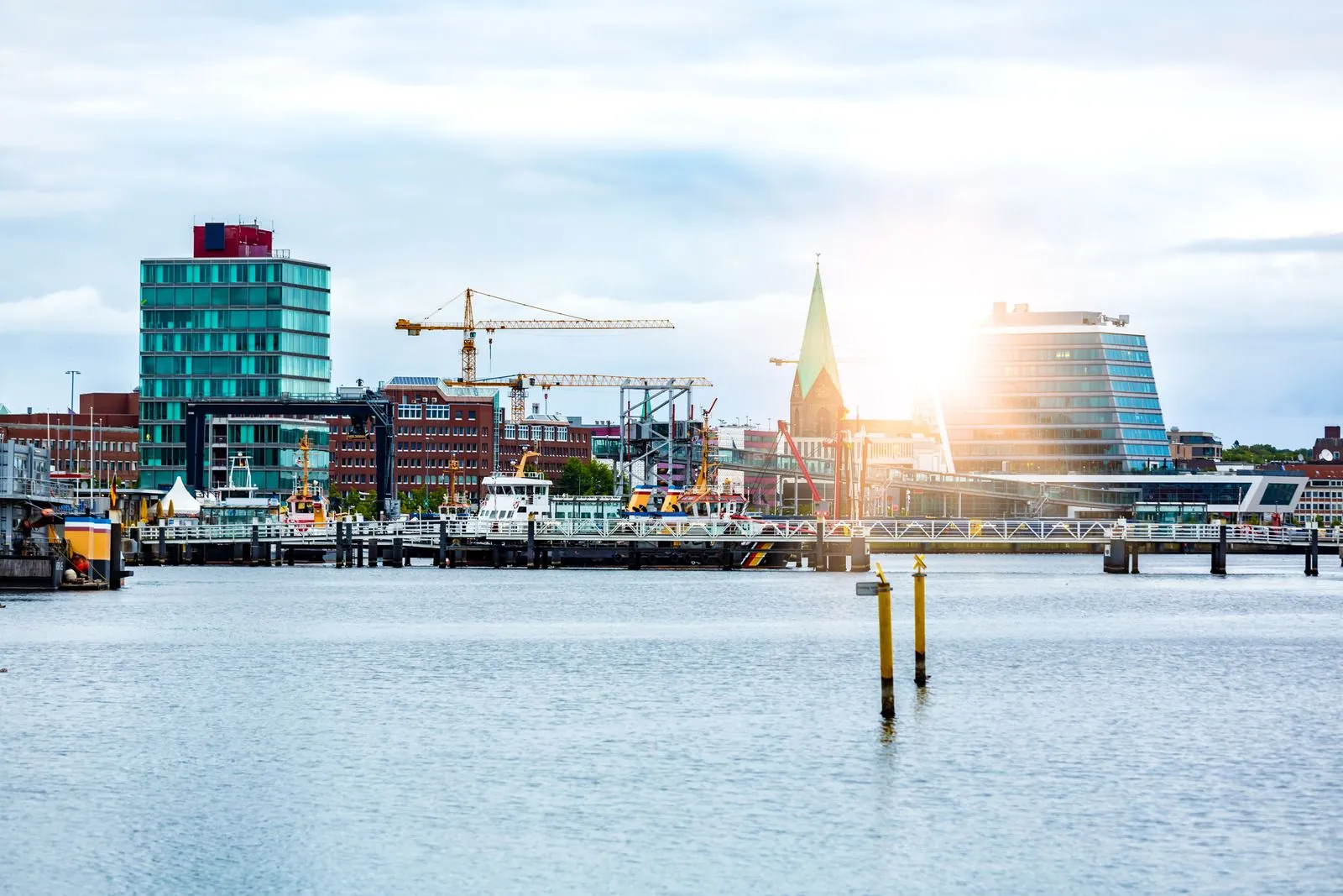 Ruhiges Hafenwasser in Kiel, dahinter eine Kaikante mit Glasfassaden, Hafenkränen und einem Kirchturm im warmen Gegenlicht